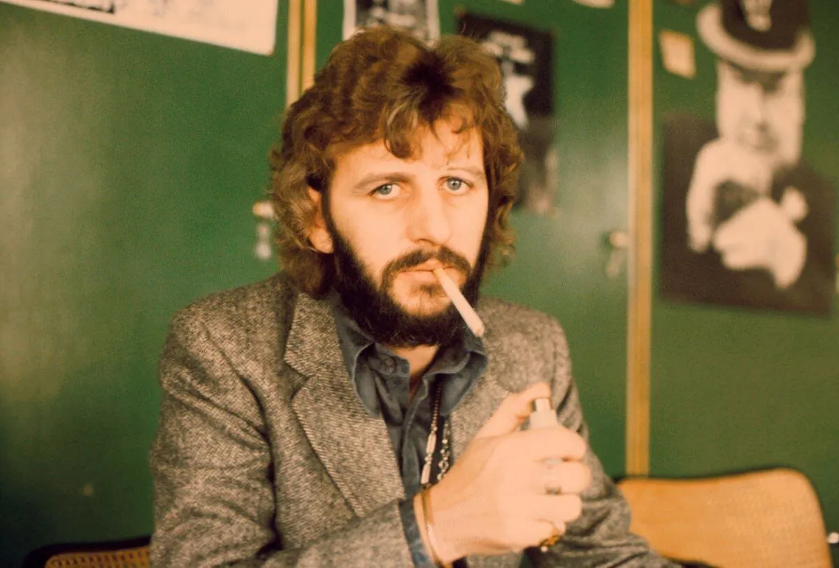 The Beatles' Ringo Starr sits in front of a green wall and lights a cigarette.