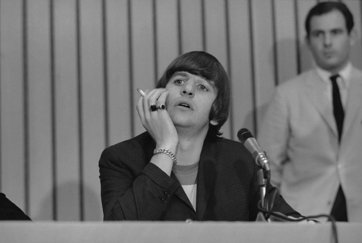 A black and white picture of Ringo Starr sitting in front of a microphone with his chin resting on his hand. He holds a cigarette between his fingers.