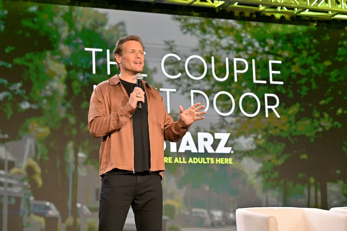 Sam Heughan standing on stage in front of a backdrop advertising 'The Couple Next Door' on Starz