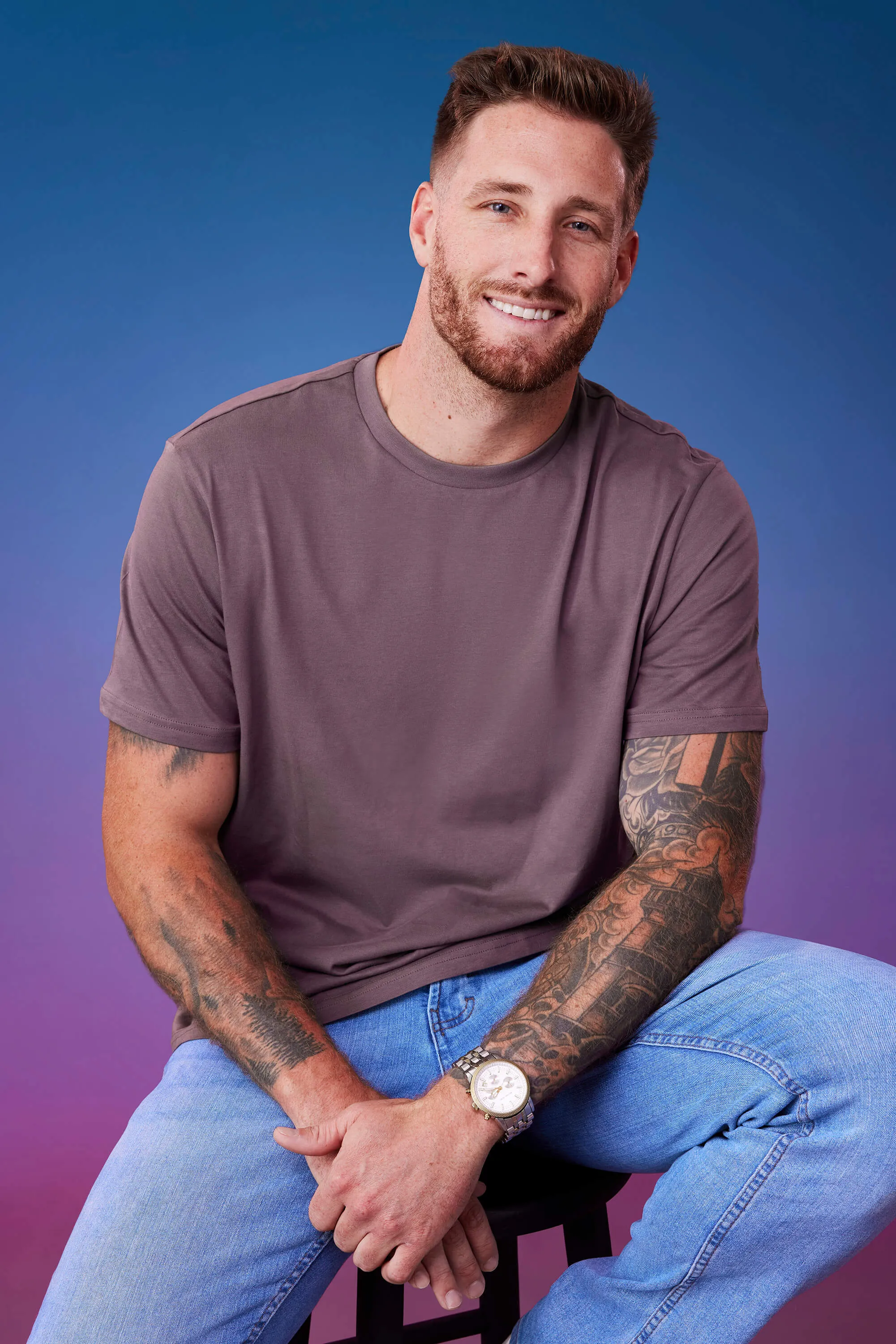 A photo of Sam McKinney from 'The Bachelorette' Season 21 sitting casually in a purple T-shirt and jeans