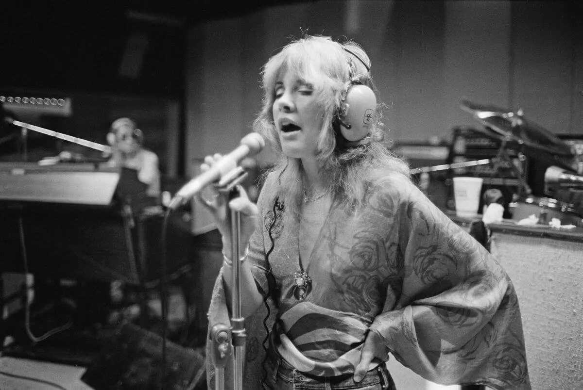 A black and white picture of Stevie Nicks wearing headphones and singing into a microphone in the studio.