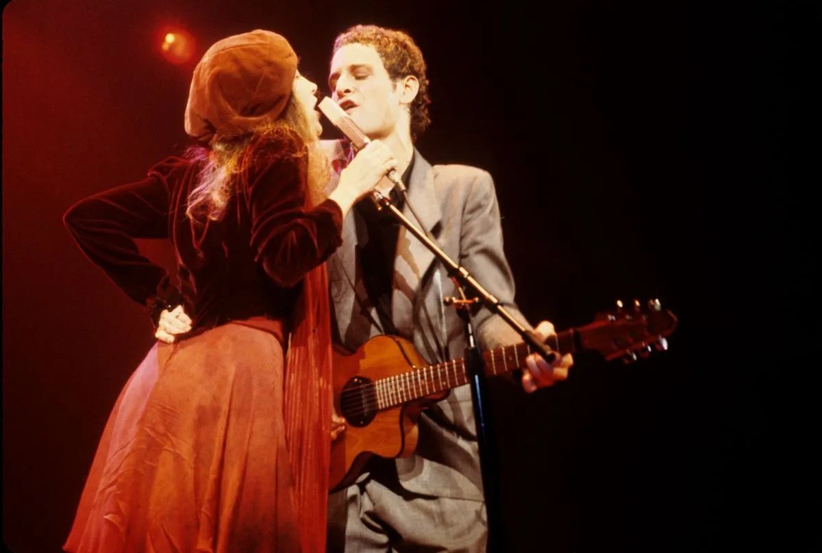 Stevie Nicks and Lindsey Buckingham sing into the same microphone. Nicks wears a beret and Buckingham plays guitar.