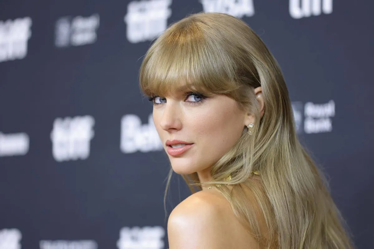 Taylor Swift stands in front of a blue background and looks over her shoulder at the camera.