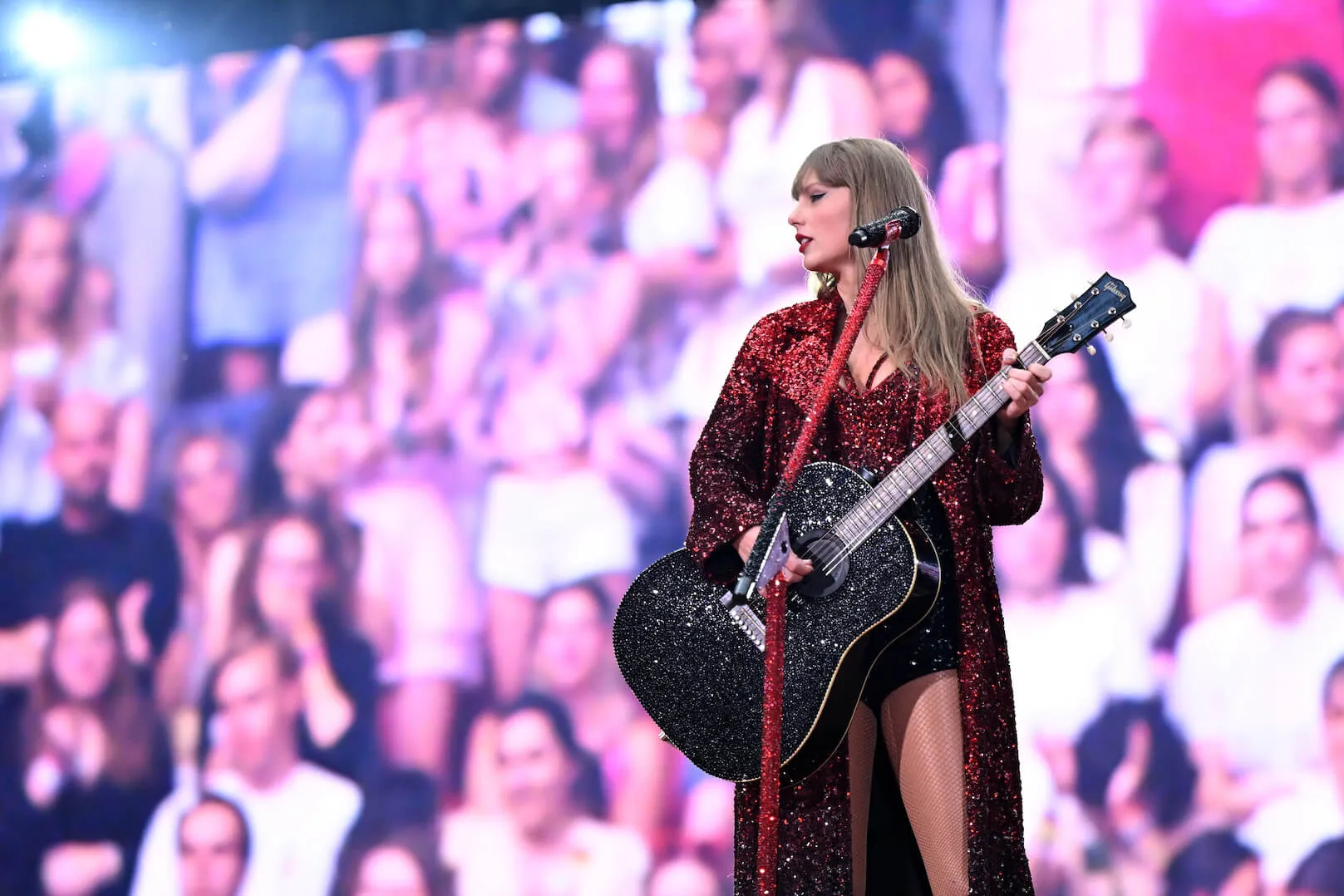 Taylor Swift holding a guitar on stage during 'The Eras Tour' against a background of people in Amsterdam