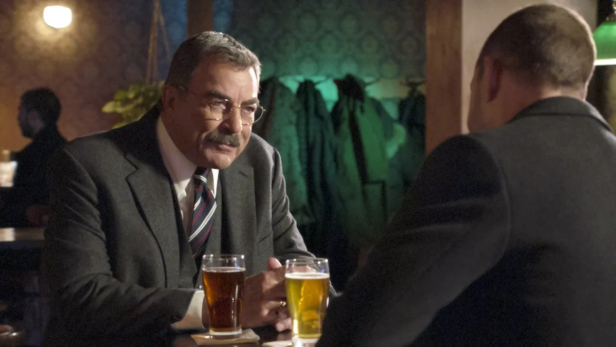Tom Selleck sitting at a table with a beer in front of him in 'Blue Bloods'