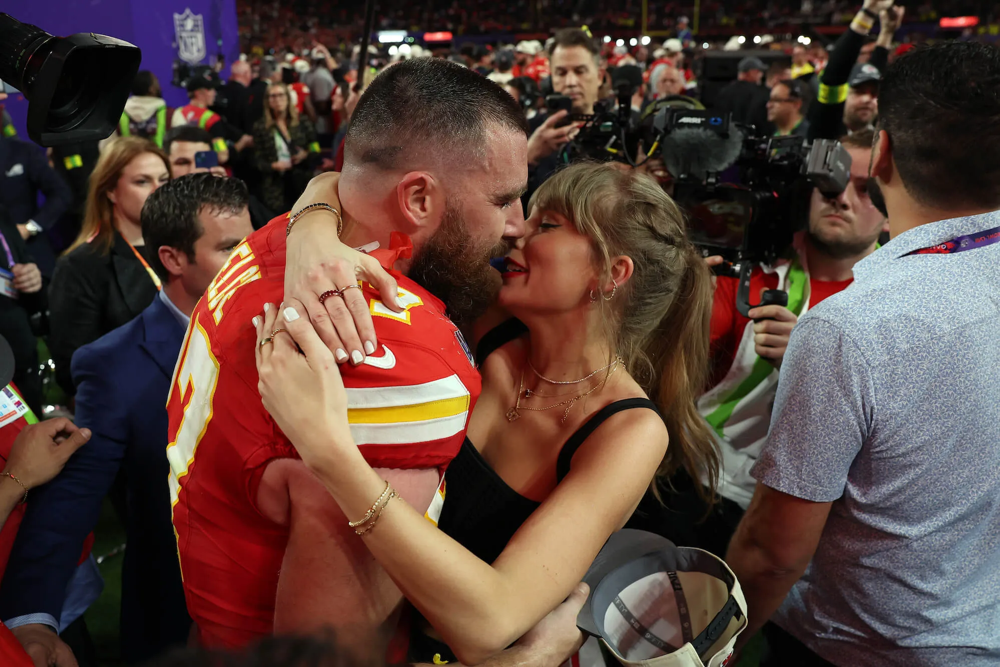 Travis Kelce and Taylor Swift embracing and kissing with a crowd surrounding them after a Kansas City Chiefs game