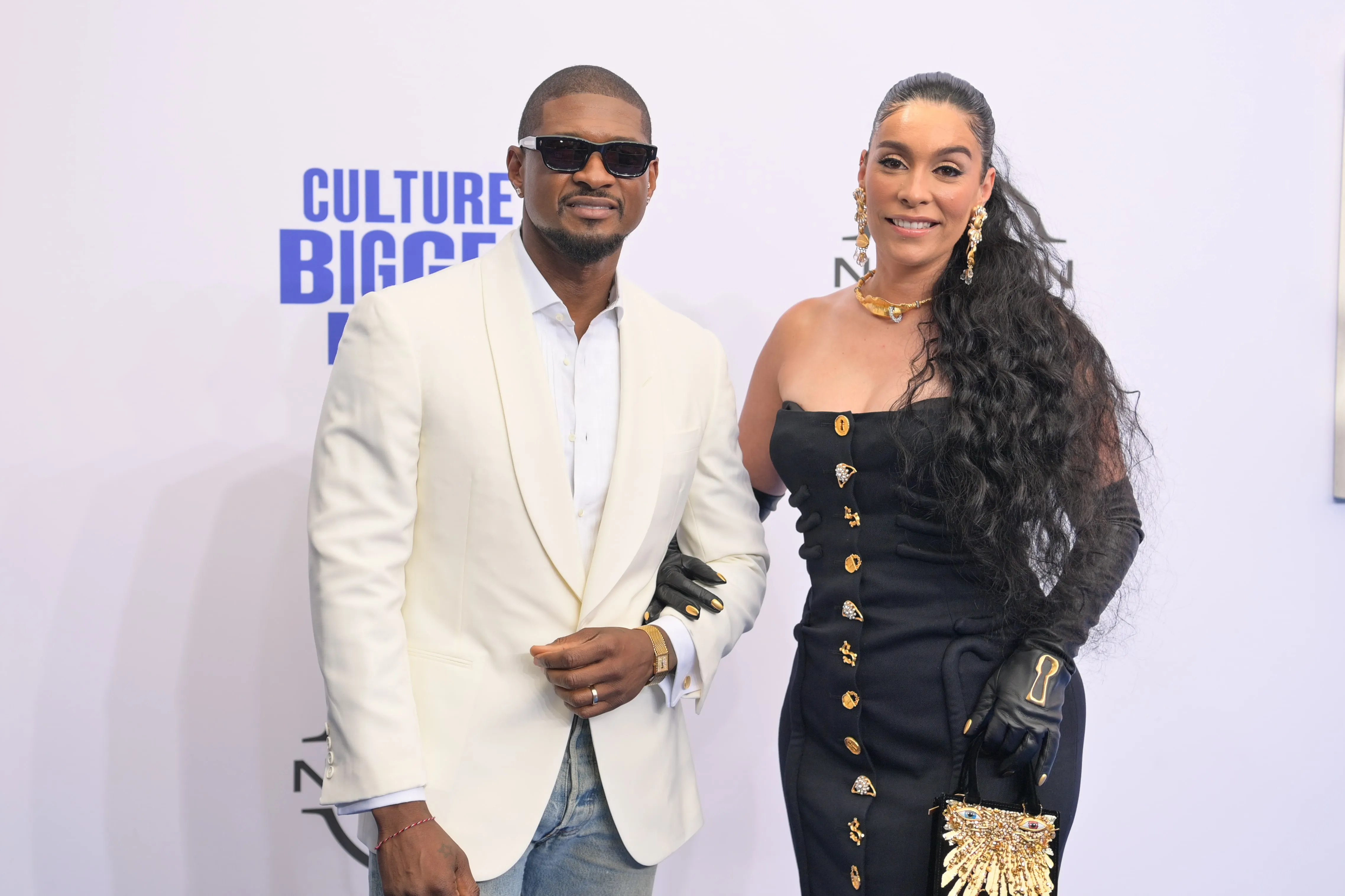 Wearing a black dress, Jennifer Goicoechea and her husband Usher smile for cameras on a red carpet