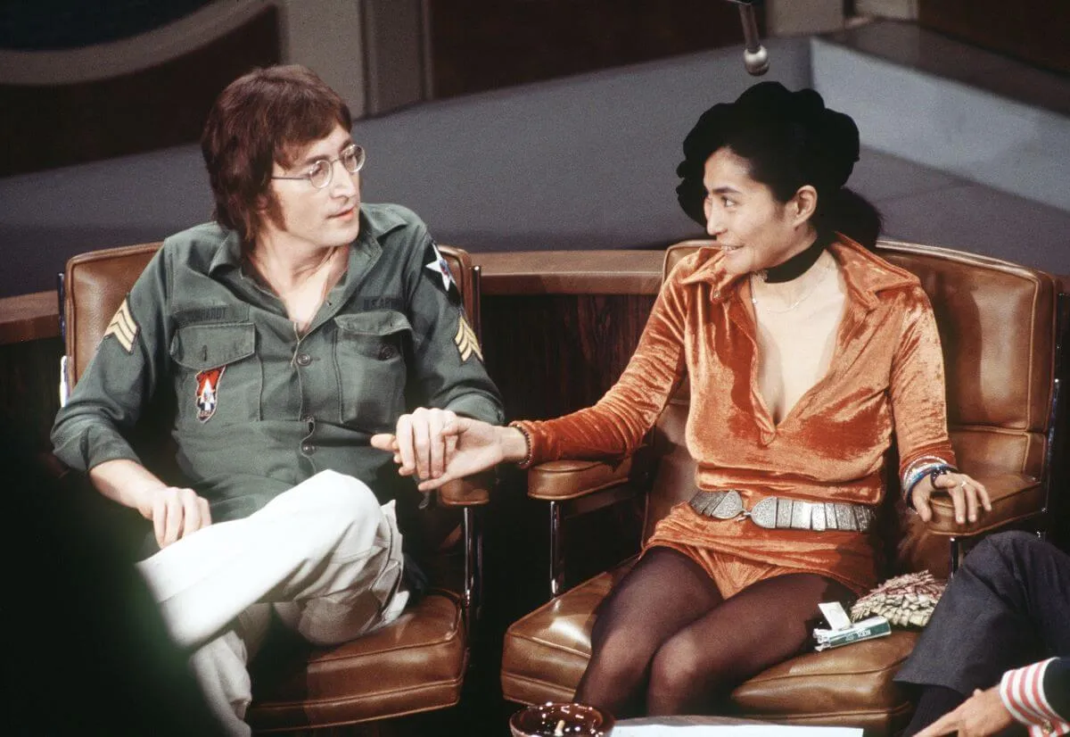 John Lennon and Yoko Ono sit next to each other and hold hands. He wears a green shirt with patches and she wears an orange romper with a hat.