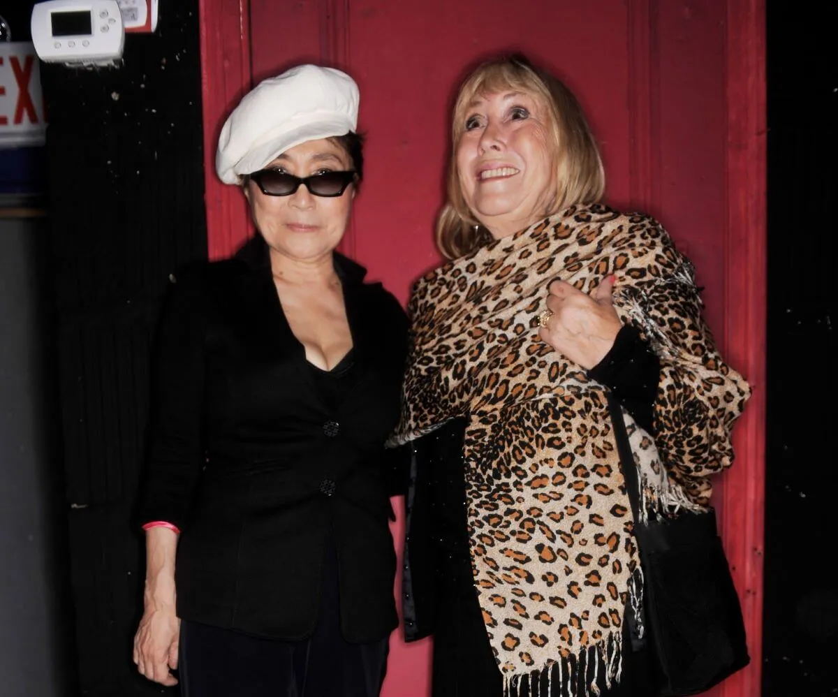 Yoko Ono wears a white hat, black sunglasses, and a black shirt and stands with Cynthia Lennon, who wears a leopard print scarf.