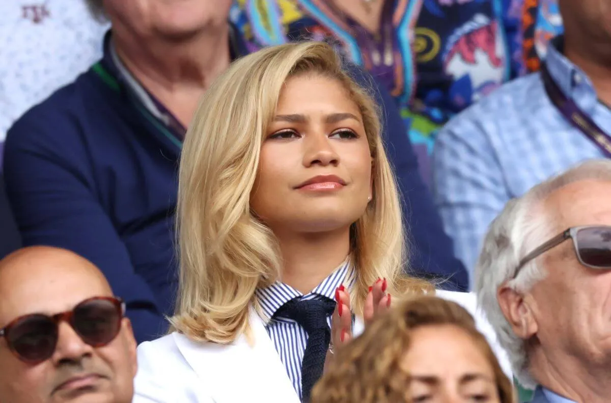 Zendaya sits in the crowd at Wimbledon while wearing a white suit jacket and tie. She claps.