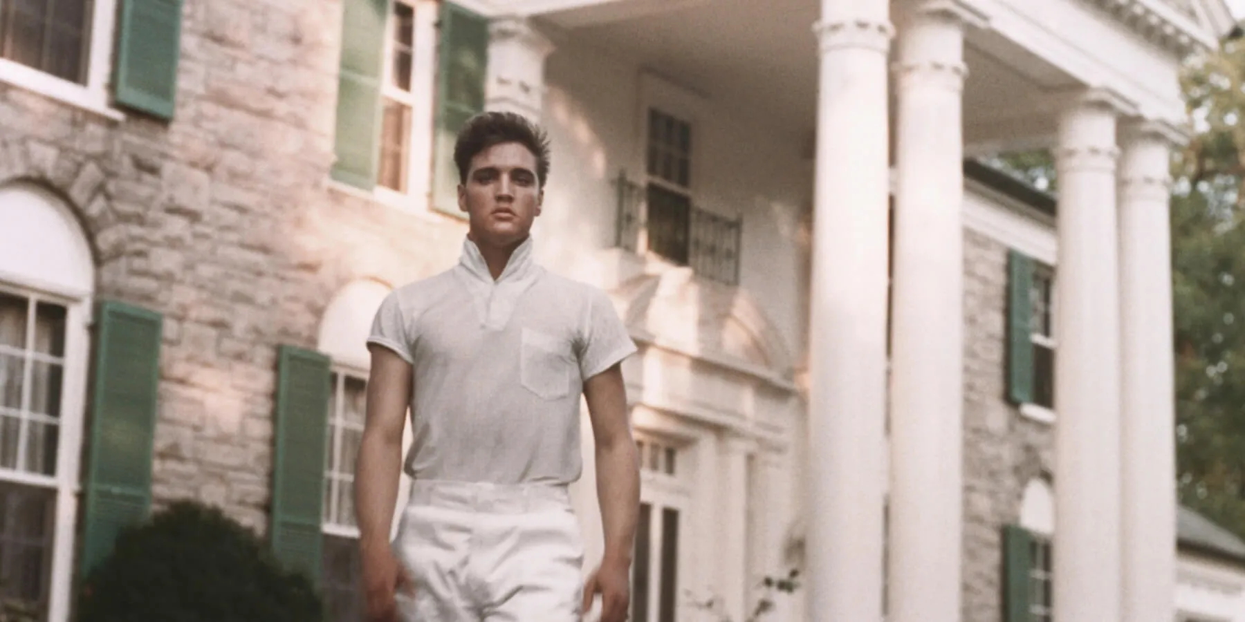 Elvis Presley in a white shirt and pants in front of Graceland