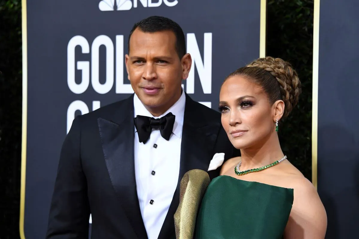 Alex Rodriguez wears a tuxedo and stands with Jennifer Lopez, who wears a green and gold dress. They stand in front of a sign for the Golden Globes.