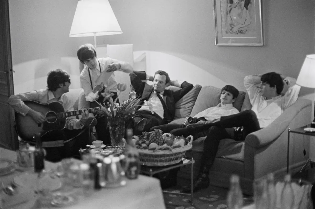 A black and white picture of Brian Epstein sitting between The Beatles on a couch. They all lounge except John Lennon, who plays guitar.