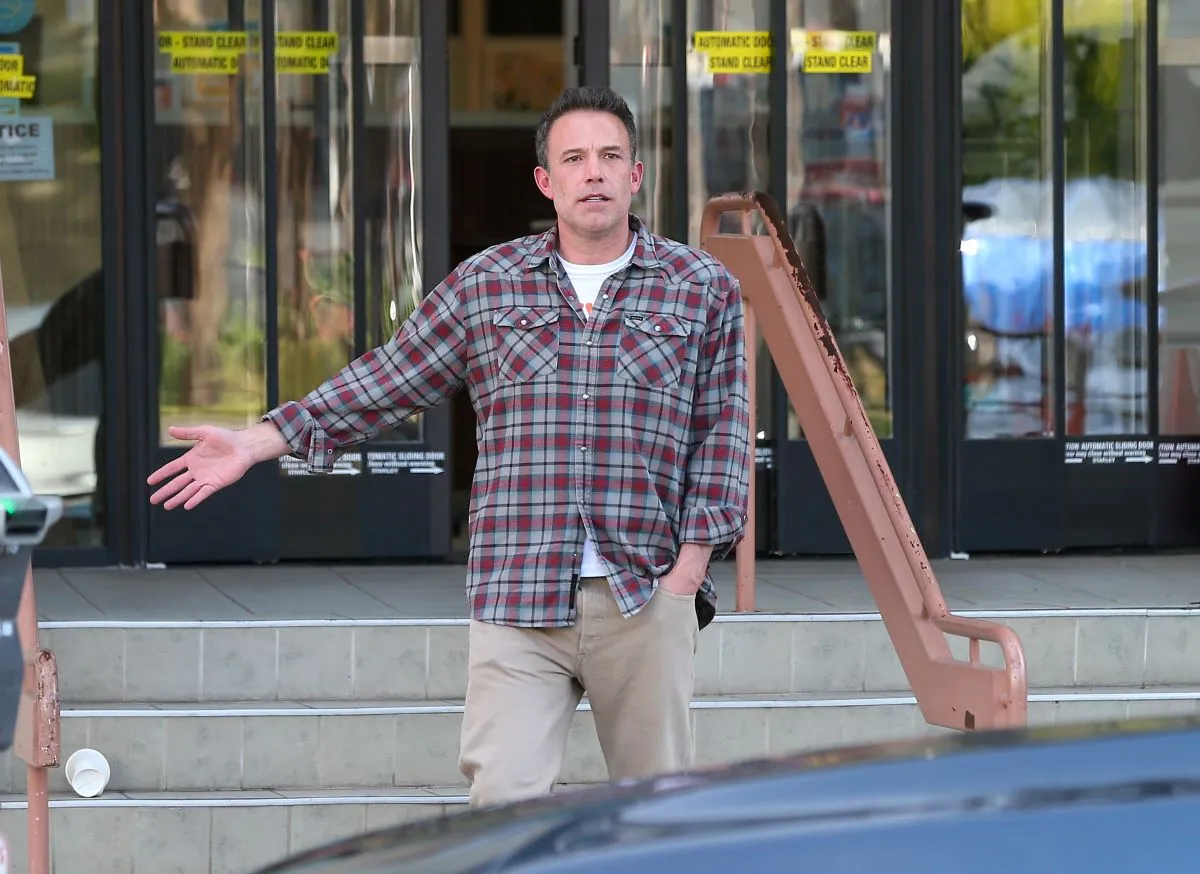 Ben Affleck wears a plaid shirt and stands in front of a building.