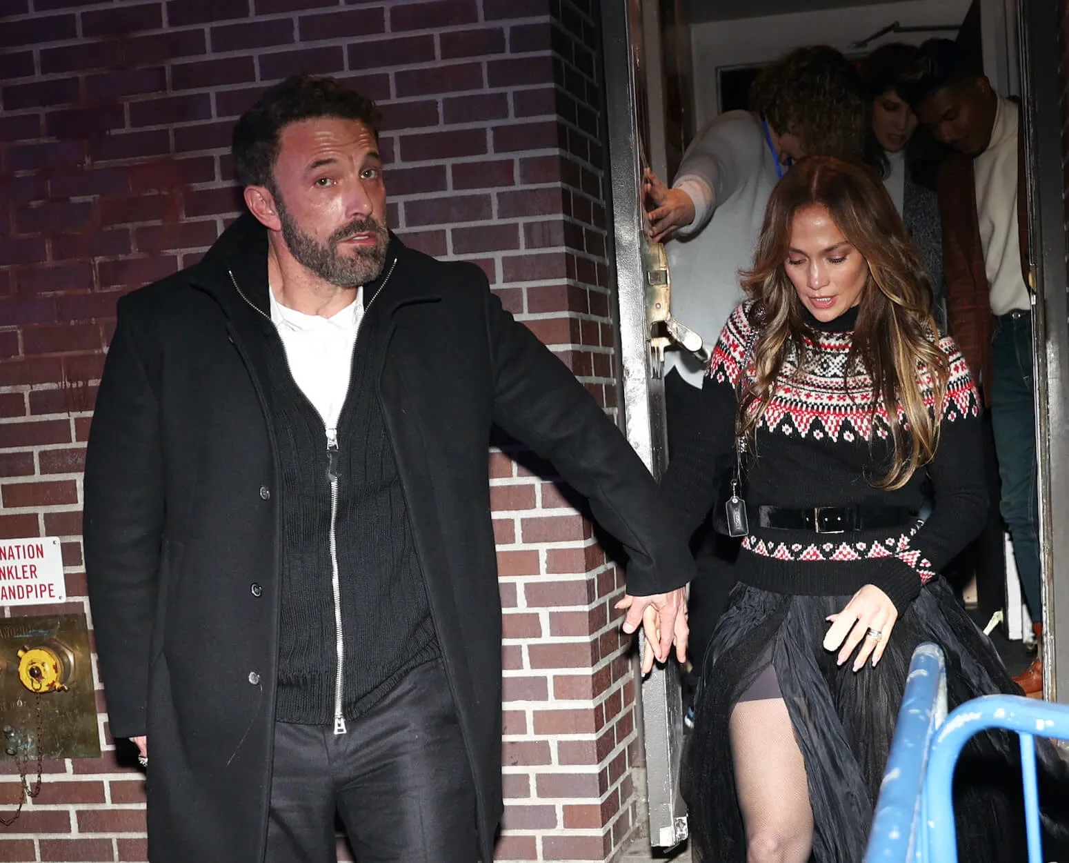 Ben Affleck holding Jennifer Lopez's hand as they walk out of a house in the dark in New York City. Affleck is looking displeased into the camera.