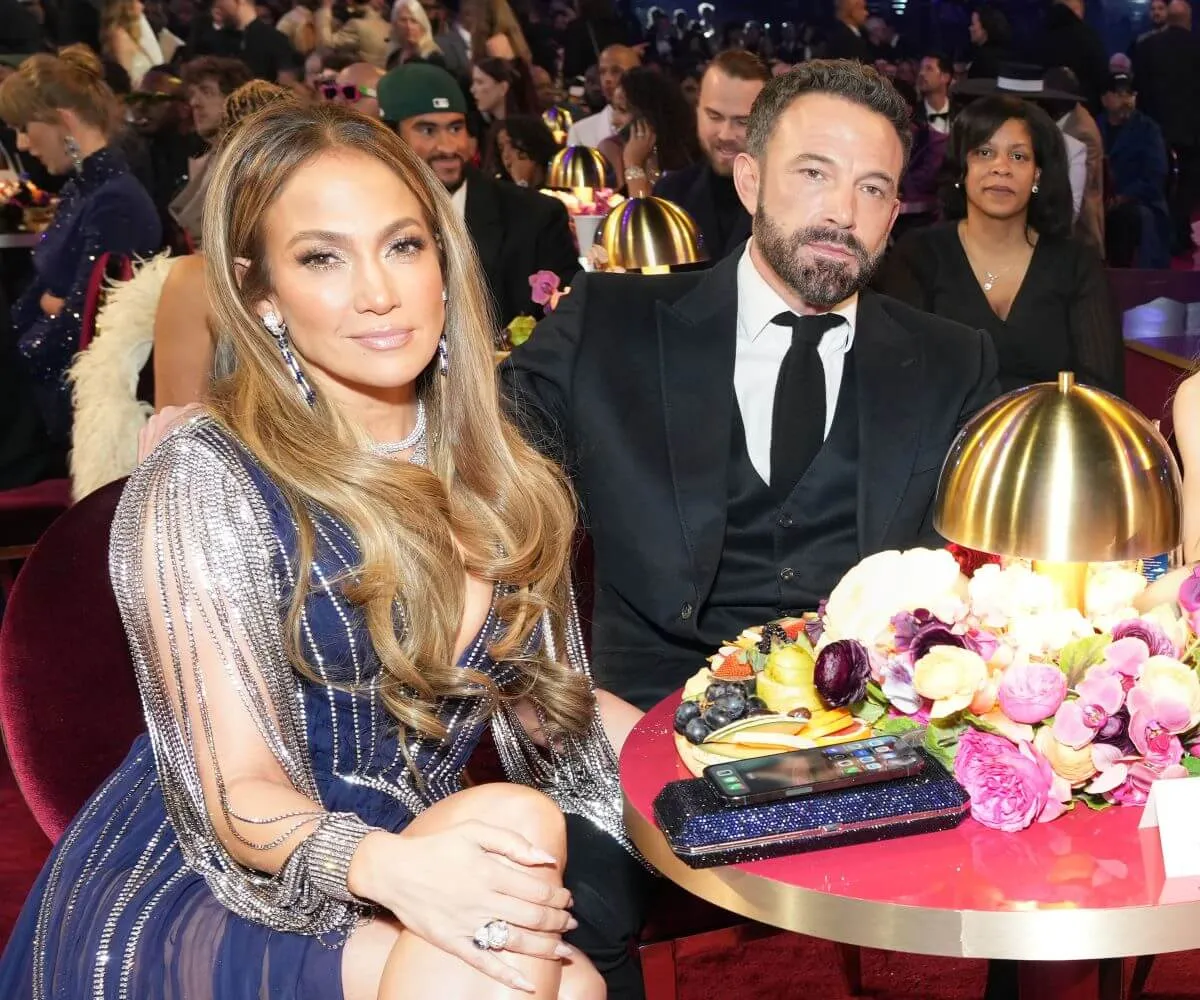 Jennifer Lopez and Ben Affleck sit at a table together. She wears a blue dress and he wears a suit.