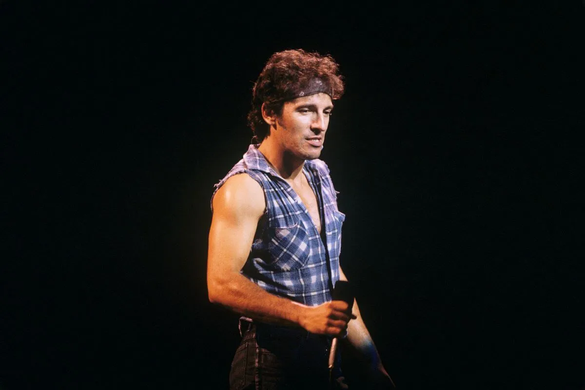 Bruce Springsteen wears a plaid shirt with the sleeves cut off and a bandanna around his head. He holds a microphone.