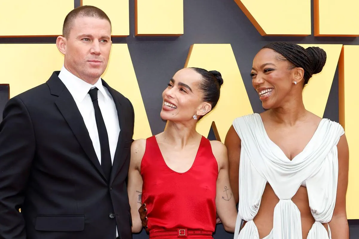 Channing Tatum, Zoe Kravitz, and Naomi Ackie stand with their arms around each other. Tatum wears a suit, Kravitz wears a red dress, and Ackie wears a white dress.