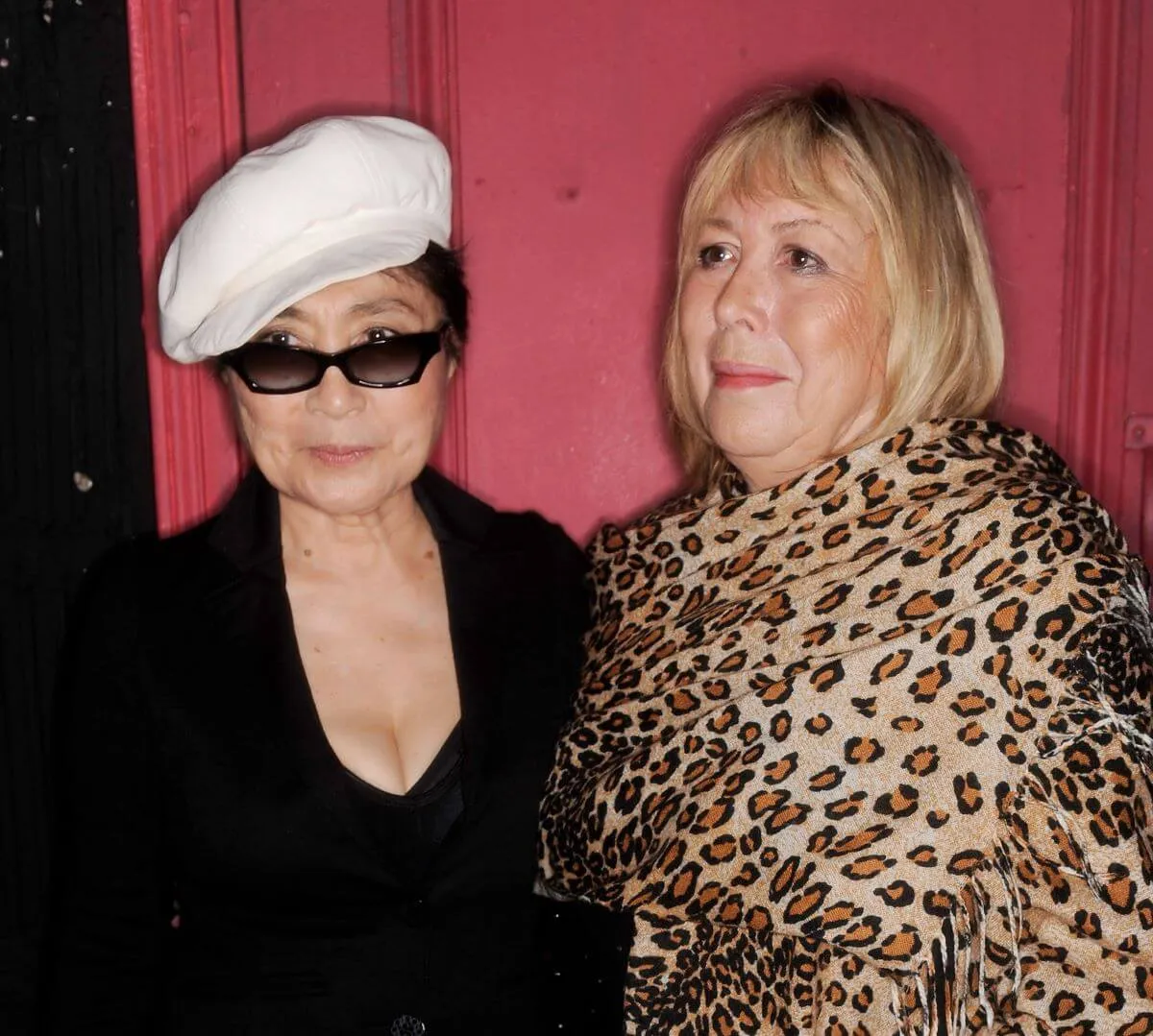 Yoko Ono and Cynthia Lennon stand in front of a red curtain. Ono wears a black shirt, white hat, and sunglasses. Lennon wears a leopard print scarf.