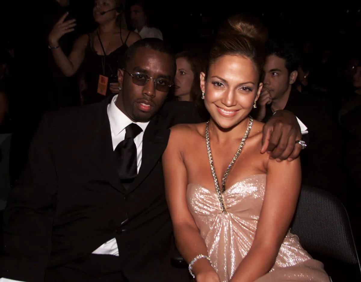 Sean 'Diddy' Combs wears a suit and sits with his arm around Jennifer Lopez's shoulder. She wears a shiny halter dress.