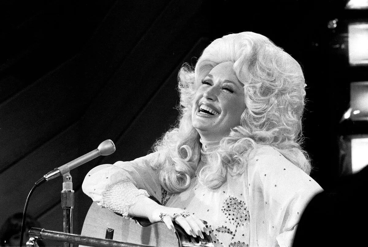 A black and white picture of Dolly Parton sitting with an acoustic guitar. She laughs.