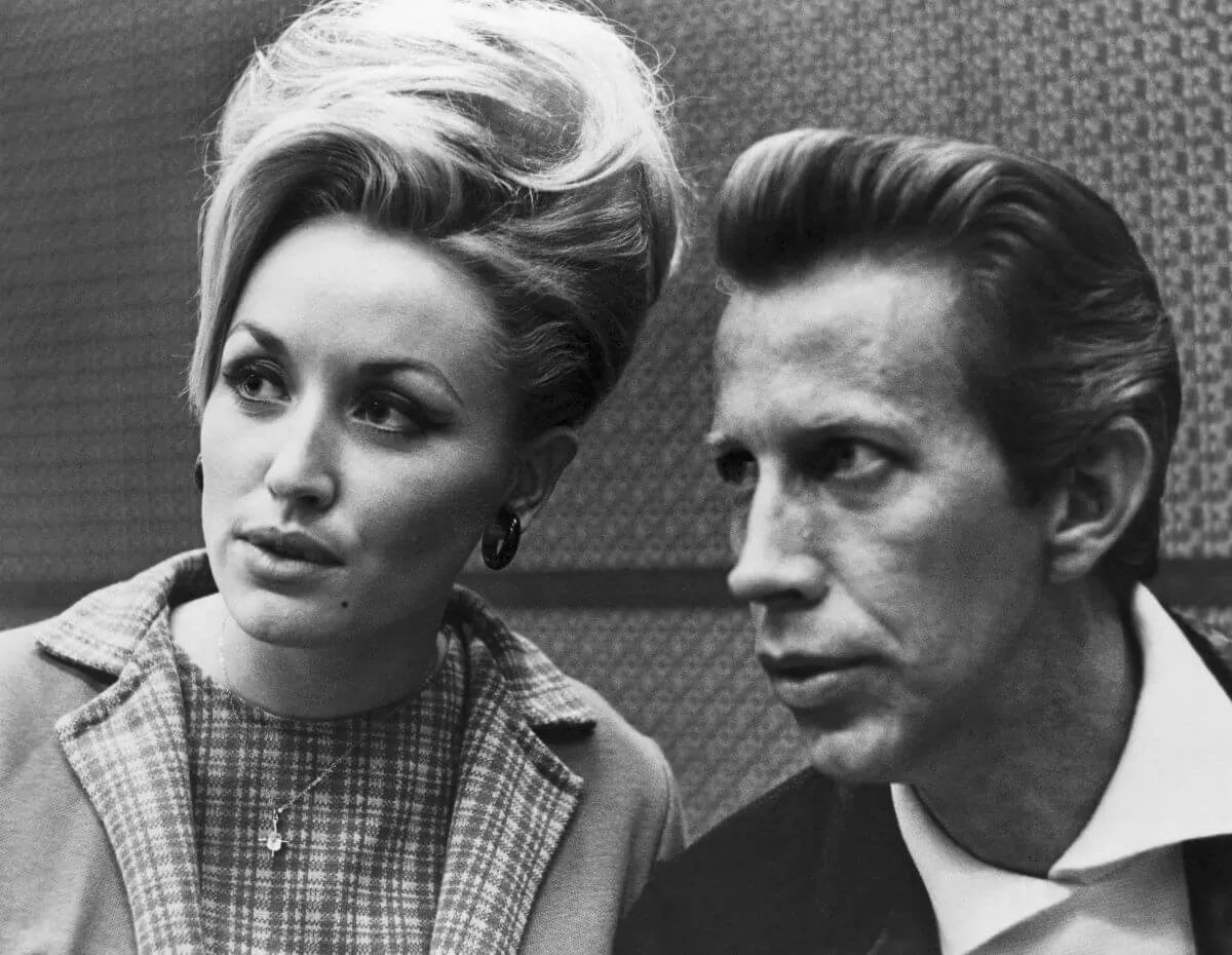 A black and white picture of Dolly Parton wearing a plaid dress and sitting with Porter Wagoner, who wears a white shirt and jacket.