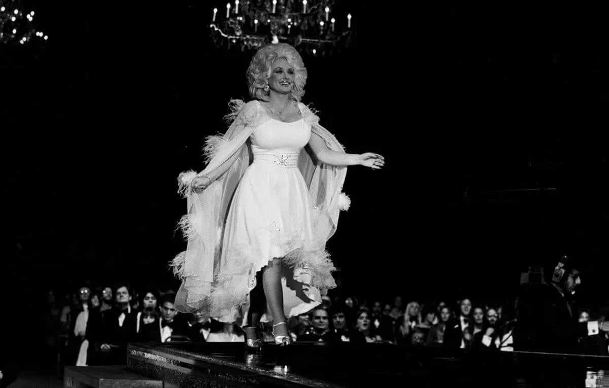 Dolly Parton walks across a stage in a dress and cape.