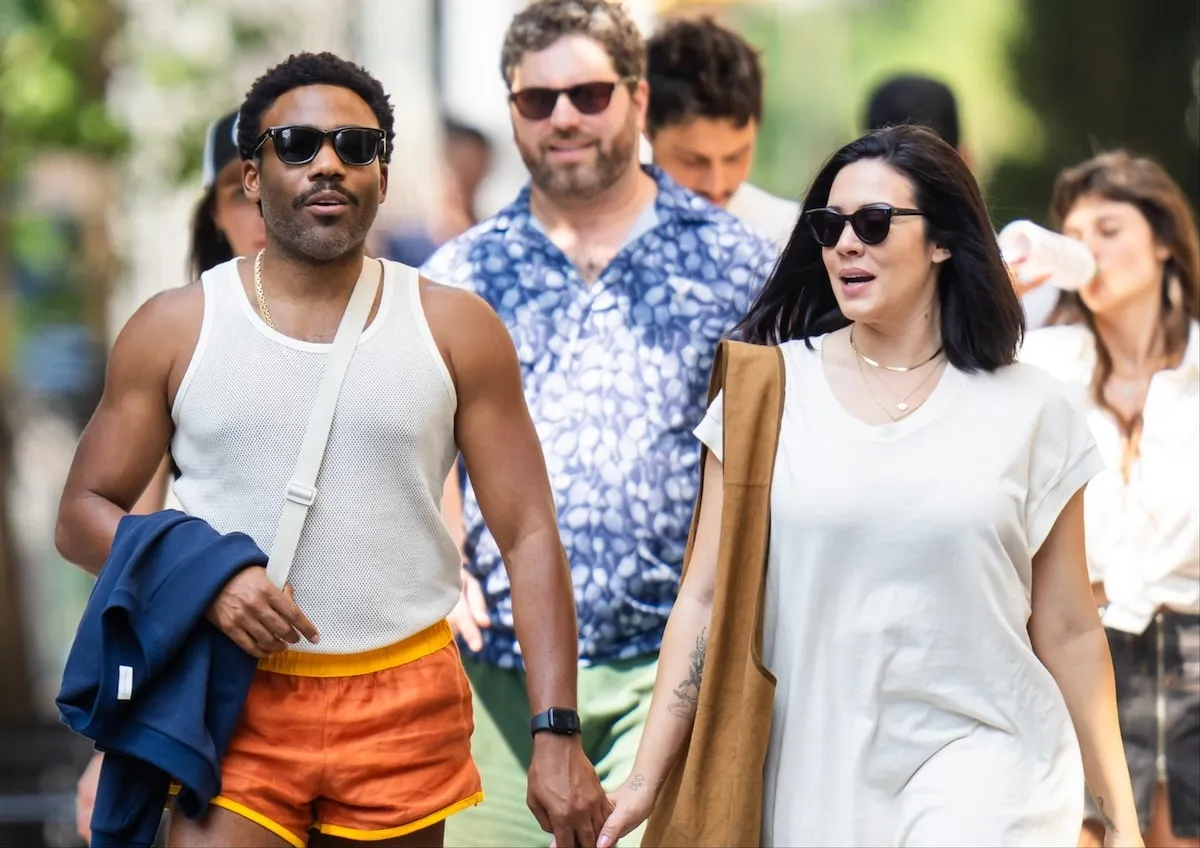 Wearing a white tank top and orange shorts, Donald Glover walks down an NYC street holding hands with his wife Michelle White, wearing a white dress