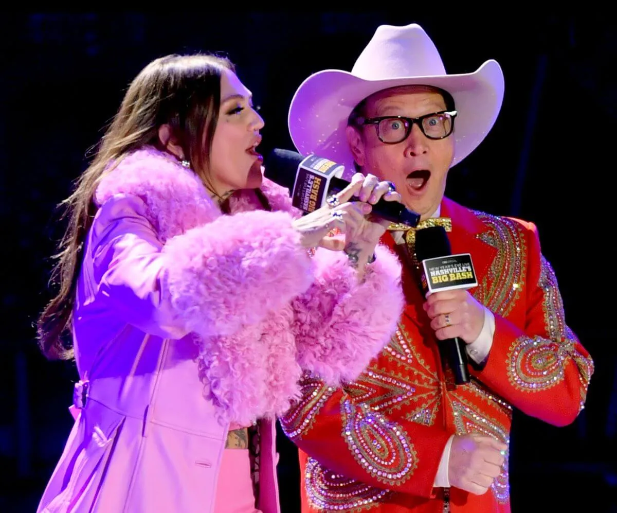 Elle King wears a pink coat with fur sleeves and stands with Rob Schneider, who wears a red suit and a cowboy hat. He also holds a microphone.