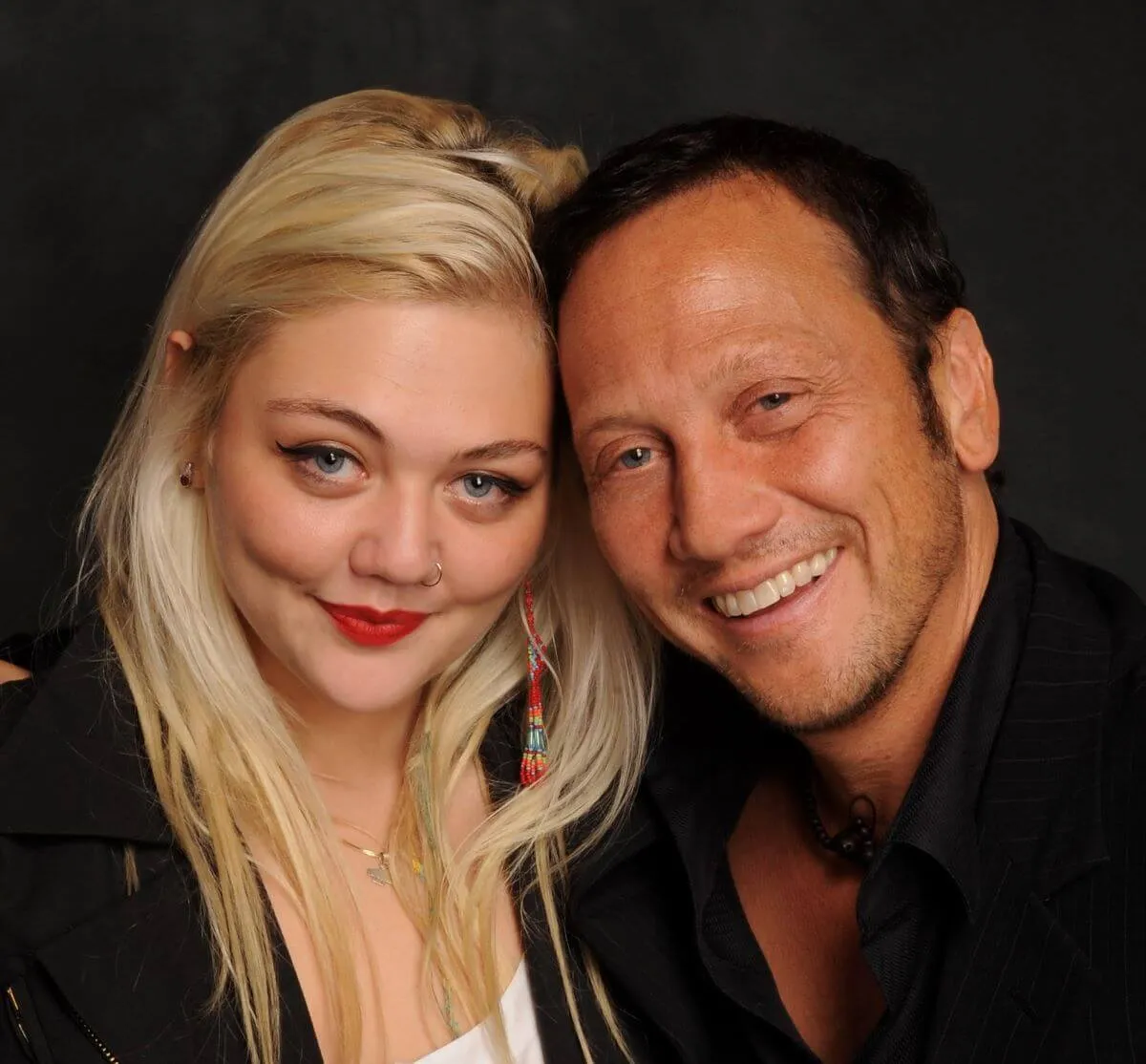 Elle King and Rob Schneider smile together. They both wear black and she has red lipstick on.