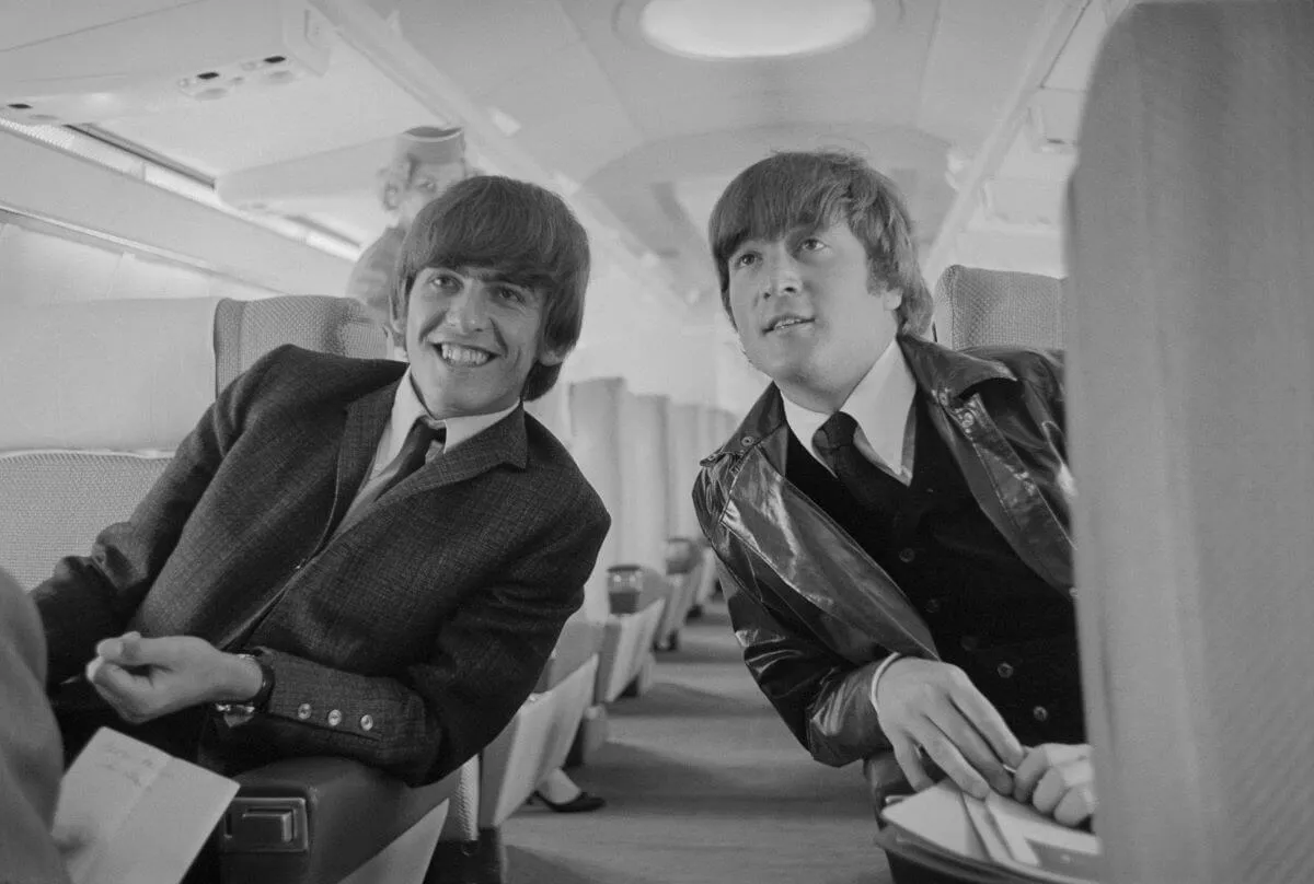 A black and white picture of George Harrison and John Lennon leaning towards each other from opposite sides of an airplane aisle.