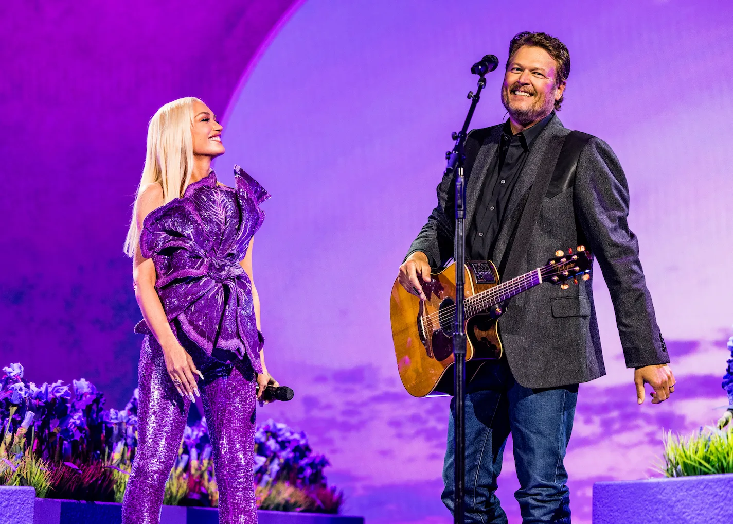 Gwen Stefani dressed in a purple dress standing on stage next to Blake Shelton in a suit. They're standing against a purple backdrop at the59th Academy of Country Music Awards