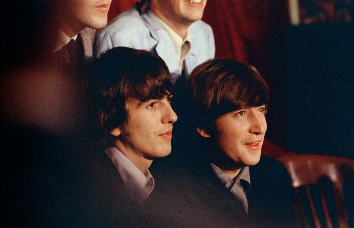 George Harrison and John Lennon sit next to each other. Paul McCartney and Ringo Starr sit behind them, and the bottom halves of their faces are visible.