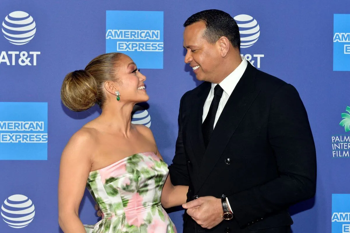 Jennifer Lopez and Alex Rodriguez stand in front of a blue background. She wears a strapless dress in a floral pattern and he wears a suit.