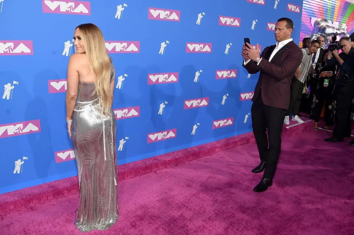 Jennifer Lopez wears a silver dress and poses for photographers. Alex Rodriguez takes a picture of her with his phone.