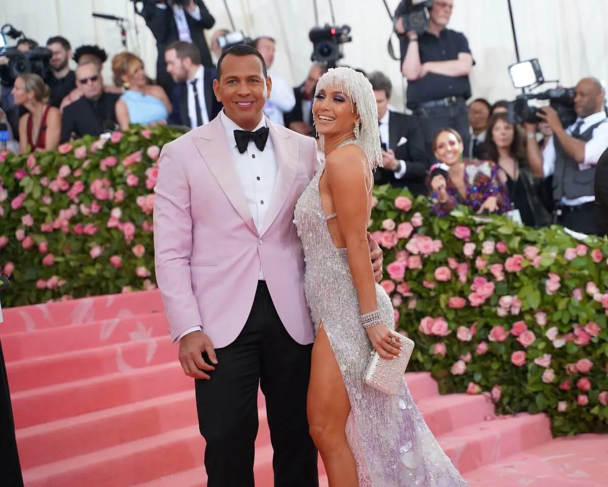 Alex Rodriguez and Jennifer Lopez stand on the steps of the Met Gala. He wears a pink suit jacket and she wears a silver dress and matching wig.