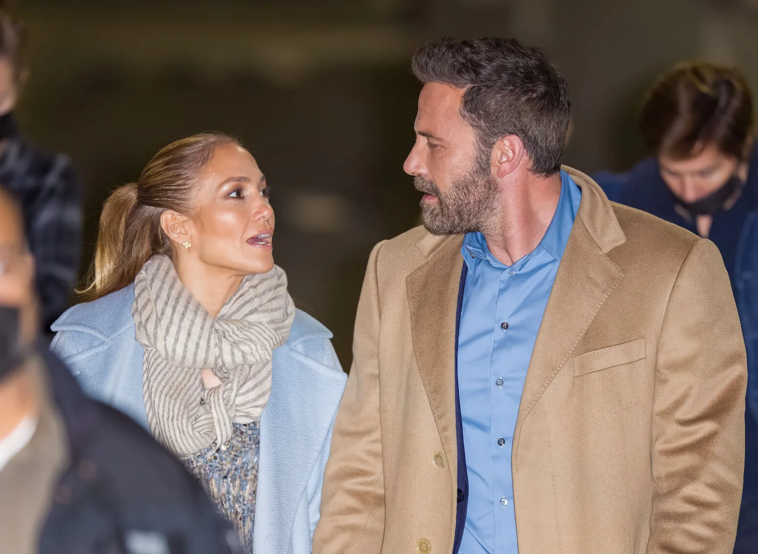 Jennifer Lopez in a blue coat and Ben Affleck in a beige coat looking at each other and talking while walking in a crowd