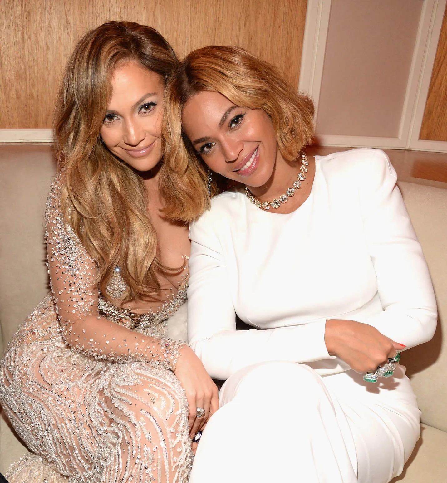 Jennifer Lopez and Beyoncé sitting on a couch and leaning into each other as they smile for the camera. They're at the 2015 Vanity Fair Oscar Party.