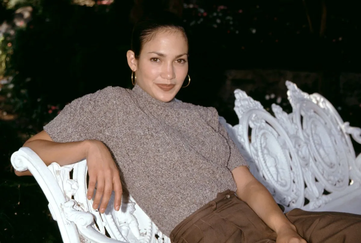 Jennifer Lopez wears a brown shirt and pants and leans on a white bench.