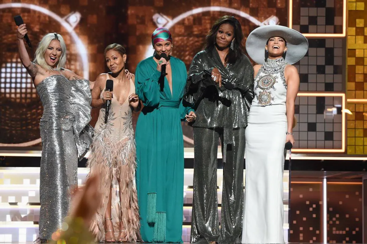 Lady Gaga, Jada Pinkett Smith, Alicia Keys, Michelle Obama, and Jennifer Lopez stand in a line together on the stage at the Grammys. Keys  holds a microphone to her mouth.