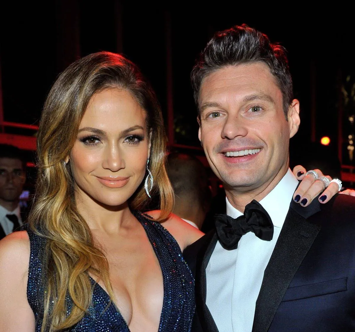 Jennifer Lopez wears a blue dress and stands with her arm around Ryan Seacrest's shoulders. He wears a tuxedo.