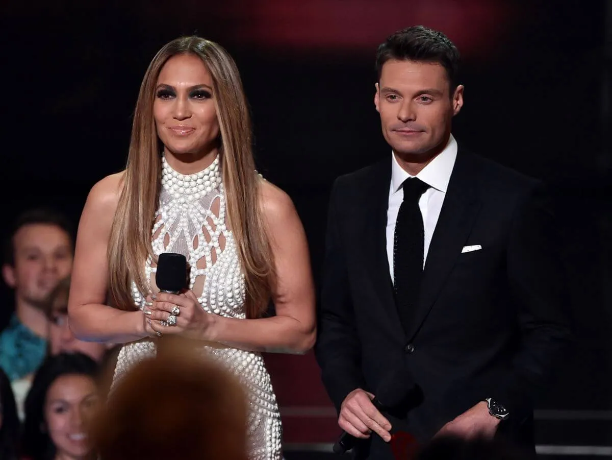 Jennifer Lopez wears a white beaded dress and stands next to Ryan Seacrest, who wears a black suit.