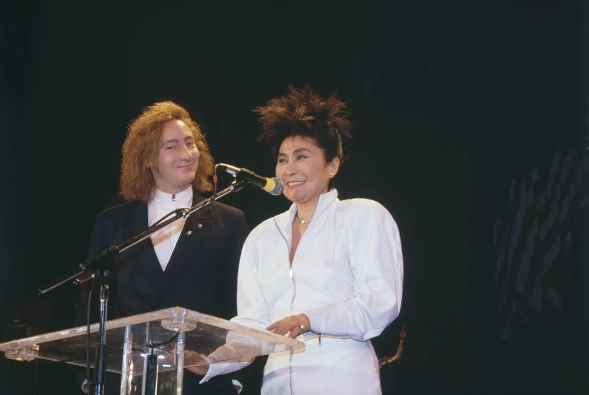 Julian Lennon and Yoko Ono stand in front of a podium. Julian wears a suit and Ono wears a white jacket.