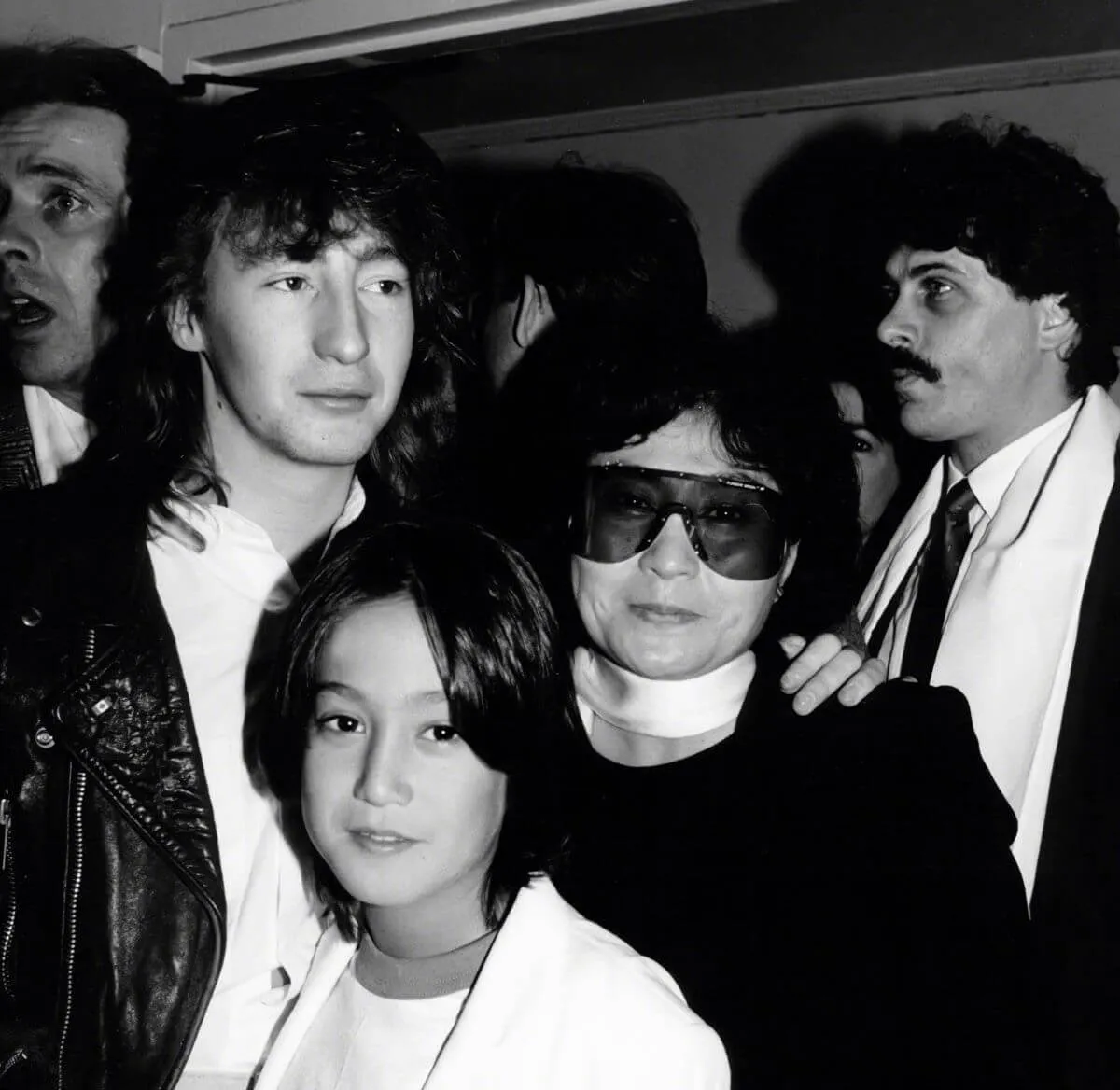 A black and white picture of Julian Lennon standing behind Yoko Ono and Sean Lennon. Ono wears sunglasses.