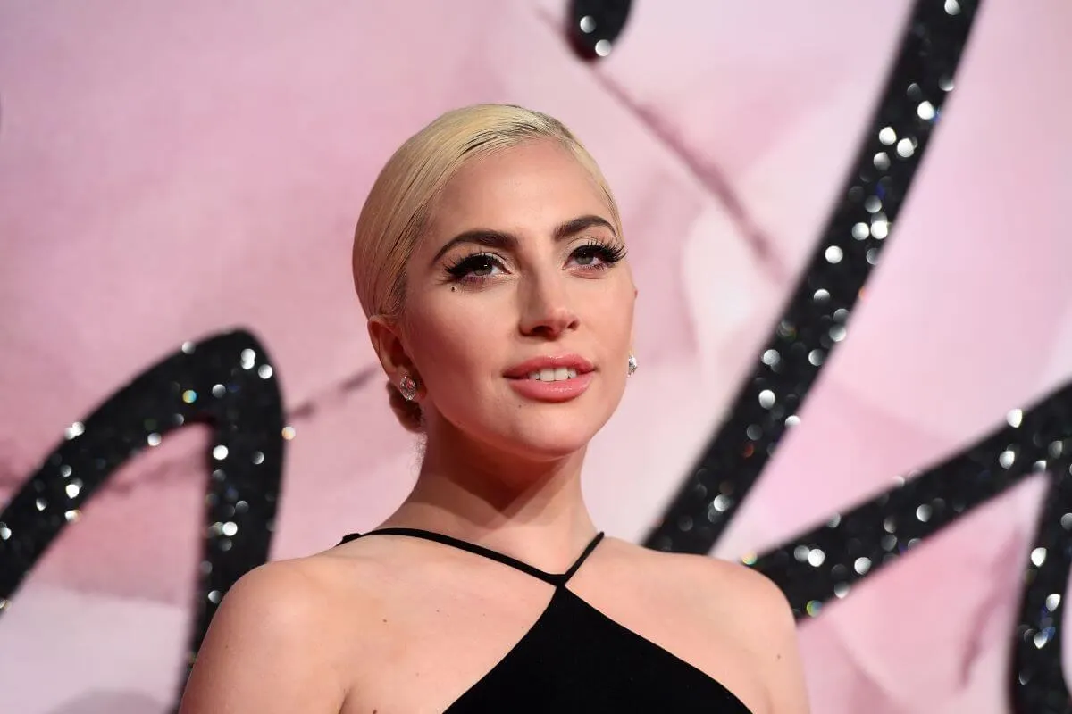 Lady Gaga wears a black dress and stands in front of a pink background.