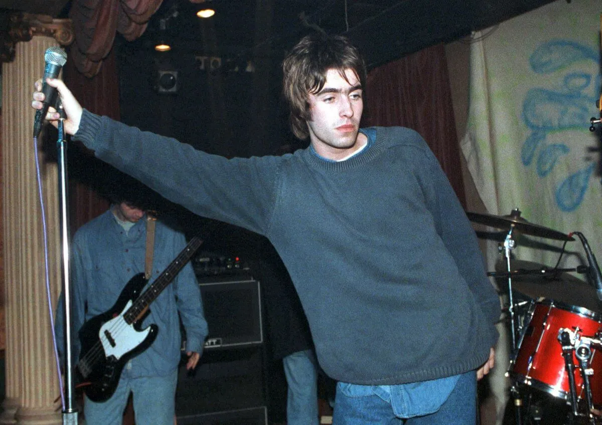 Liam Gallagher wears a sweater and holds a microphone out to the side.