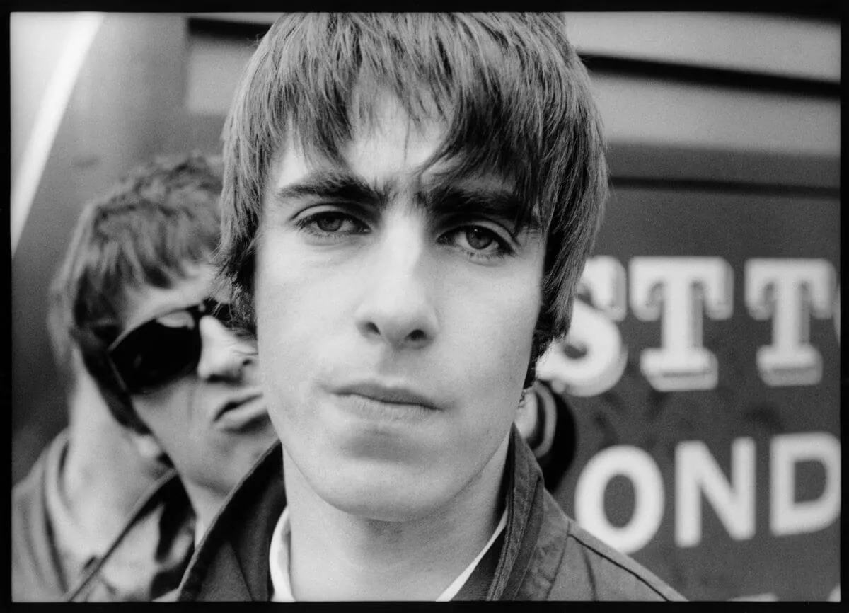 A black and white picture of Liam Gallagher looking at the camera. Noel Gallagher stands behind him wearing sunglasses.