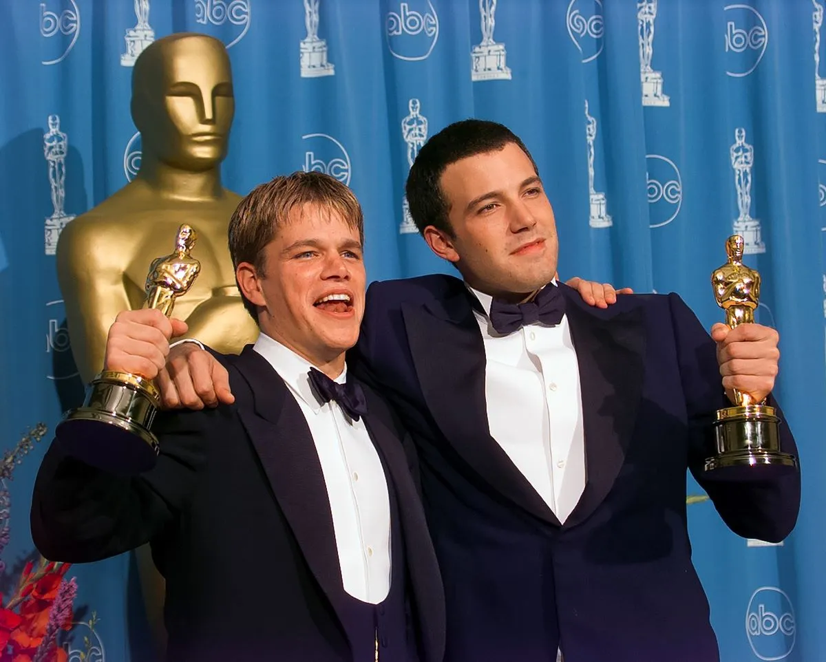 Matt Damon and Ben Affleck stand in front of a blue curtain and large Oscar statue while holding their Oscars. They have their arms around each other.