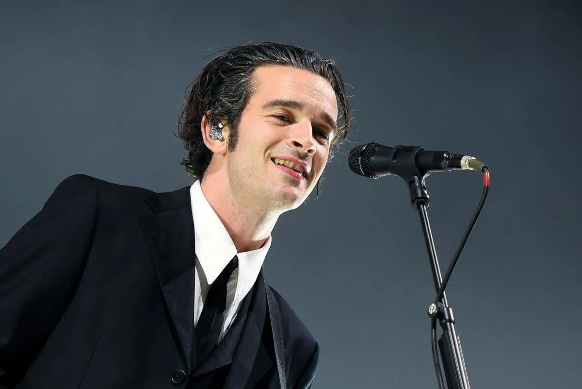 Matty Healy wears a suit and stands in front of a microphone.