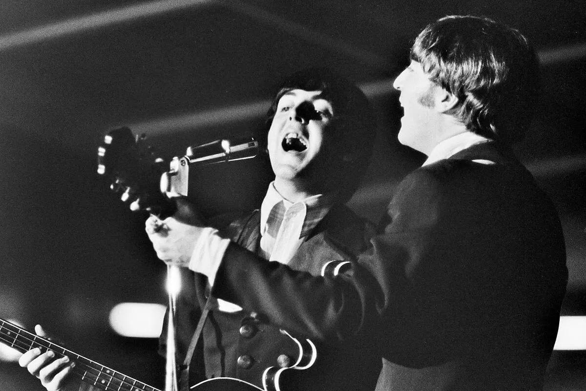 A black and white picture of Paul McCartney and John Lennon singing into the same microphone. Lennon has his back to the camera and McCartney is angled toward it.